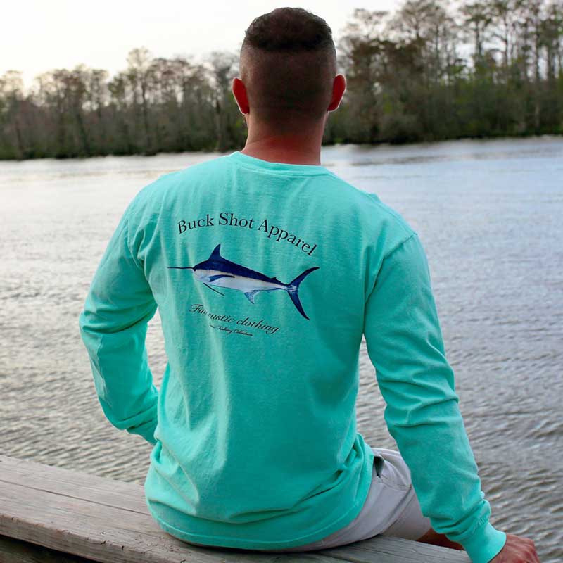 A man sitting on the dock of a lake wearing a long sleeve shirt.
