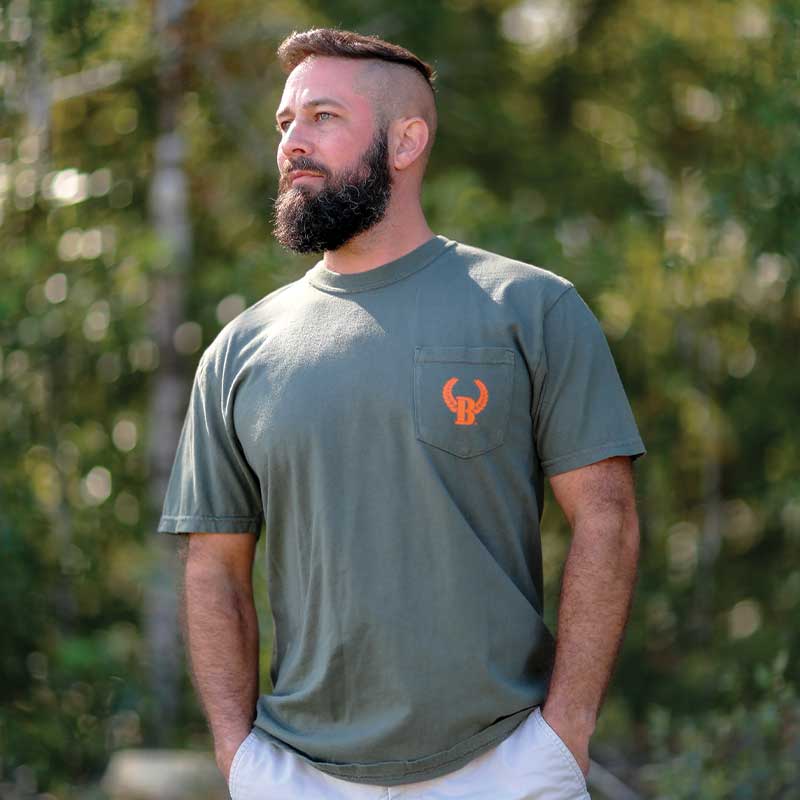 A man with a beard and a mohawk haircut stands in front of trees.