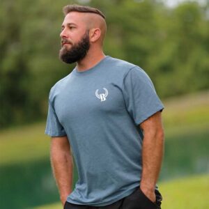 A man with a beard and a mohawk haircut stands in front of some water.