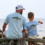 Father and son sitting on driftwood near the lake wearing matching t-shirts with Crappie graphic