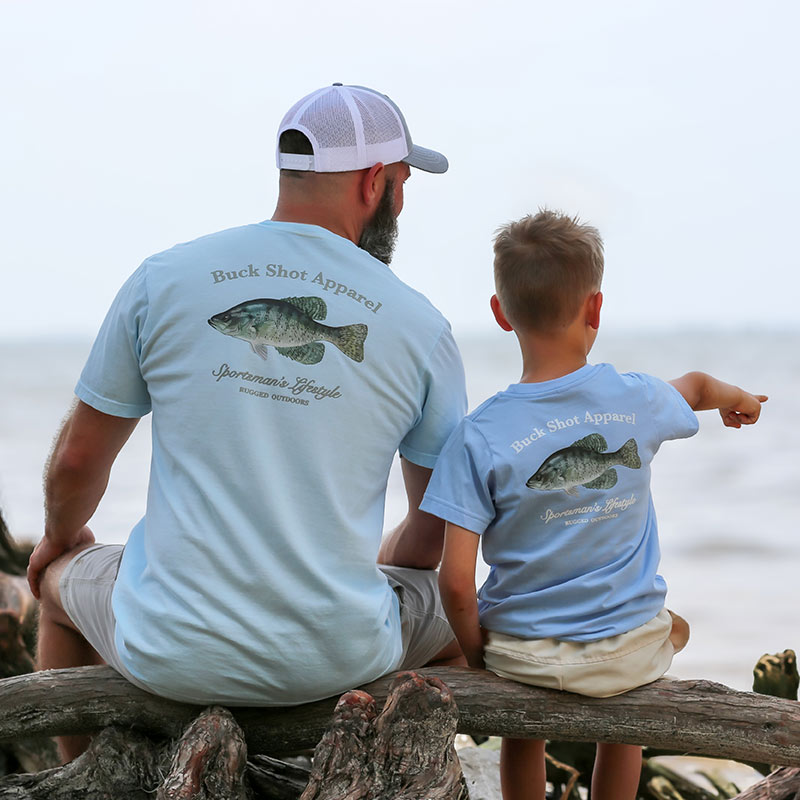 crappie father and son on driftwood by lake Father and son sitting on driftwood near the lake wearing matching t-shirts with Crappie graphic